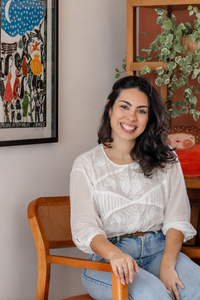 Clara Brito sentada em uma cadeira de madeira, sorrindo para a câmera, em ambiente interno com obra de arte e plantas ao fundo.