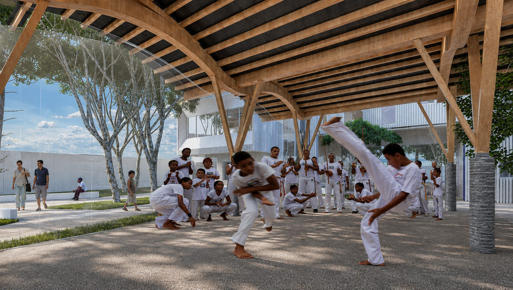 Grupo pratica capoeira em área coberta por estrutura de madeira, cercada por painéis transparentes e integrada ao pátio arborizado do centro.