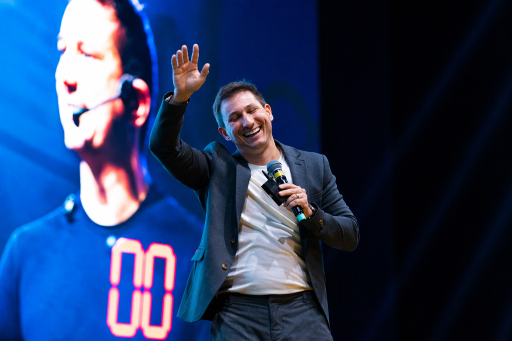 Homem sorridente em um palco, vestindo blazer preto sobre camiseta branca e segurando um microfone. Ele levanta uma das mãos enquanto fala ao público. Ao fundo, um grande telão azul exibe a imagem ampliada de uma pessoa.