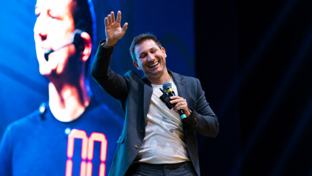 Homem sorridente em um palco, vestindo blazer preto sobre camiseta branca e segurando um microfone. Ele levanta uma das mãos enquanto fala ao público. Ao fundo, um grande telão azul exibe a imagem ampliada de uma pessoa.