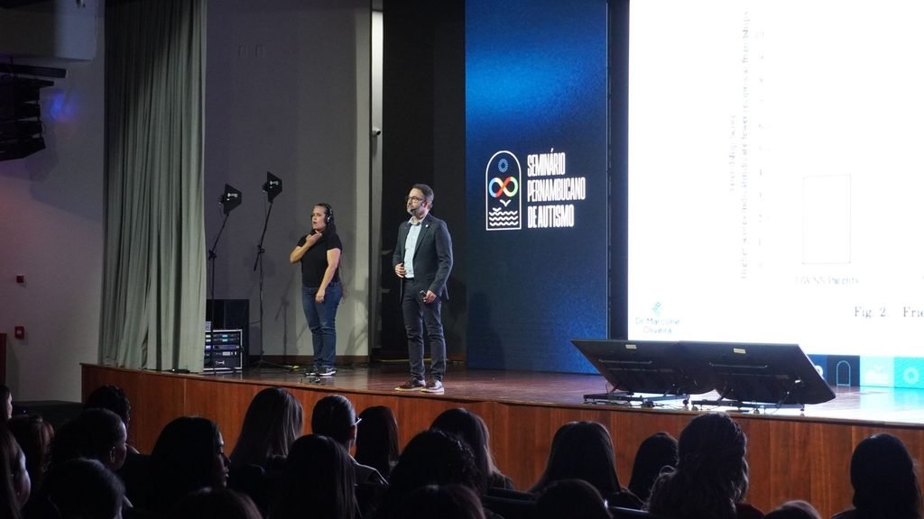 Palestrante no palco do II Seminário Pernambucano de Autismo, acompanhado por intérprete de Libras, diante de um telão com apresentação.