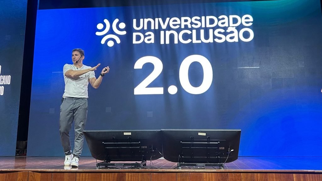 Palestrante gesticula no palco diante de um grande telão azul com a inscrição “Universidade da Inclusão 2.0”, durante o II Seminário Pernambucano de Autismo.