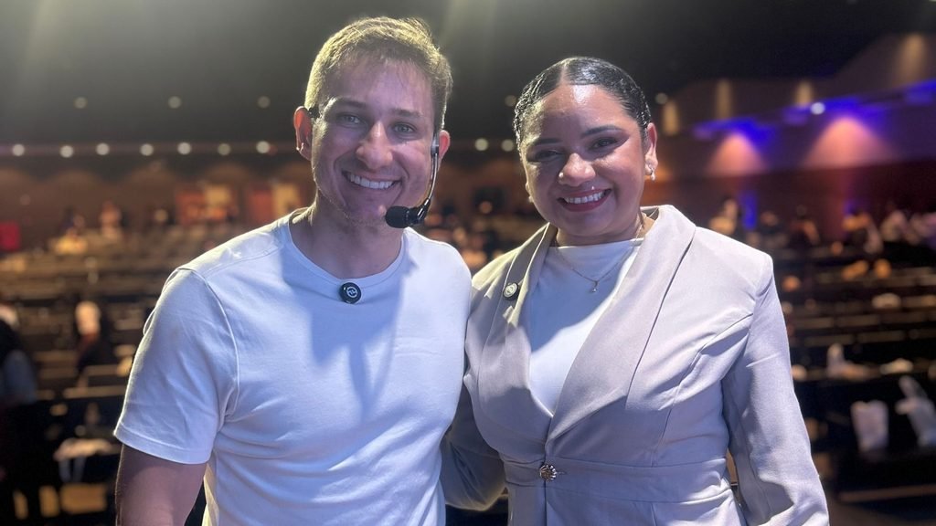 Homem e mulher sorrindo lado a lado no auditório do II Seminário Pernambucano de Autismo. O homem usa camiseta branca e microfone headset, e a mulher veste conjunto claro.