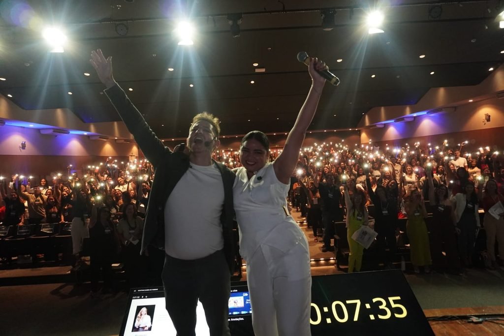 Duas pessoas no palco, de braços erguidos, diante do público do II Seminário Pernambucano de Autismo, que segura celulares com as lanternas acesas.