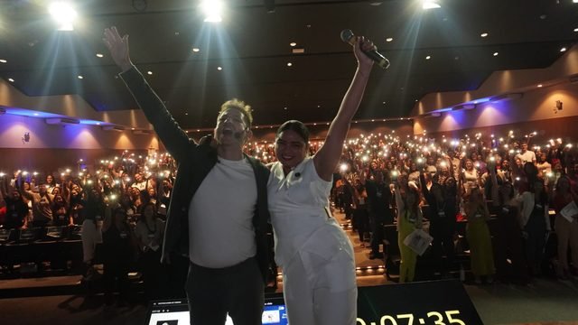 Duas pessoas no palco, de braços erguidos, diante do público do II Seminário Pernambucano de Autismo, que segura celulares com as lanternas acesas.