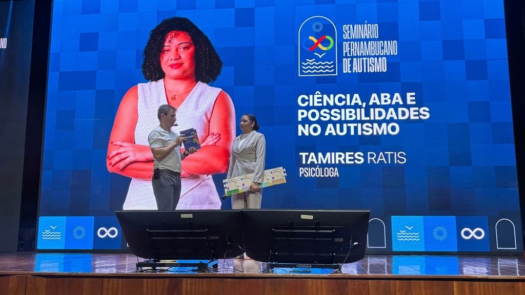 Duas pessoas no palco diante de telão que apresenta a palestra “Ciência, ABA e possibilidades no autismo”, de Tamires Ratis, durante o II Seminário Pernambucano de Autismo.
