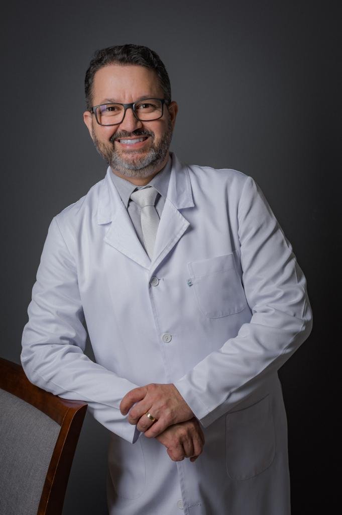 Homem sorridente, de óculos e barba curta grisalha, posa usando jaleco branco sobre camisa social e gravata. Ele está com as mãos apoiadas à frente do corpo, diante de um fundo cinza-escuro.