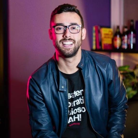Homem de pele branca, barba e cabelo curto, óculos. Veste jaqueta de couro e camisa preta. 